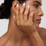 A woman with smooth skin and dark hair in a low bun touches her face, displaying the Noa Link Ring in Gold on her finger. She has natural makeup, nude-colored nails, and gazes to the side against a neutral background.