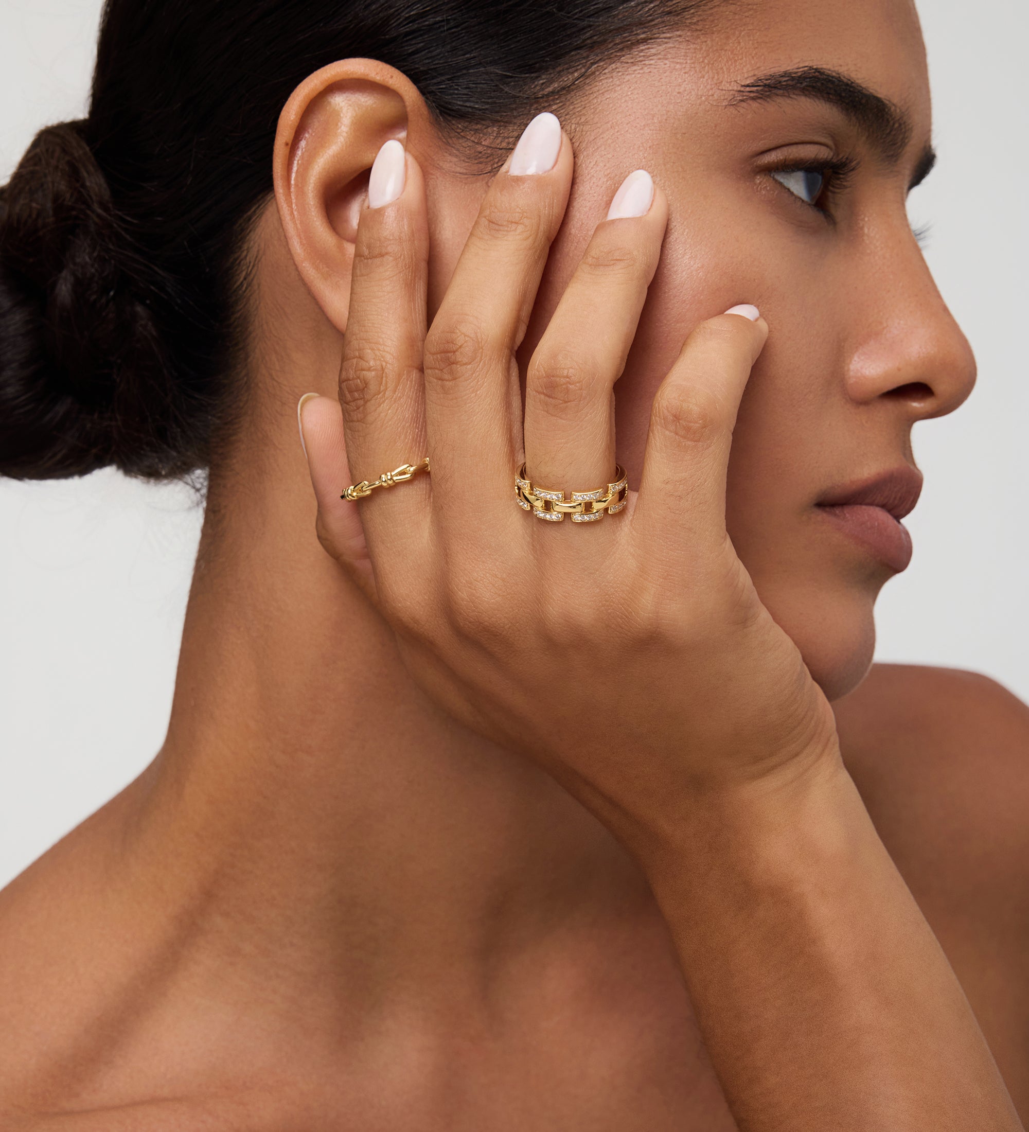 A woman with smooth skin and dark hair in a low bun touches her face, displaying the Noa Link Ring in Gold on her finger. She has natural makeup, nude-colored nails, and gazes to the side against a neutral background.