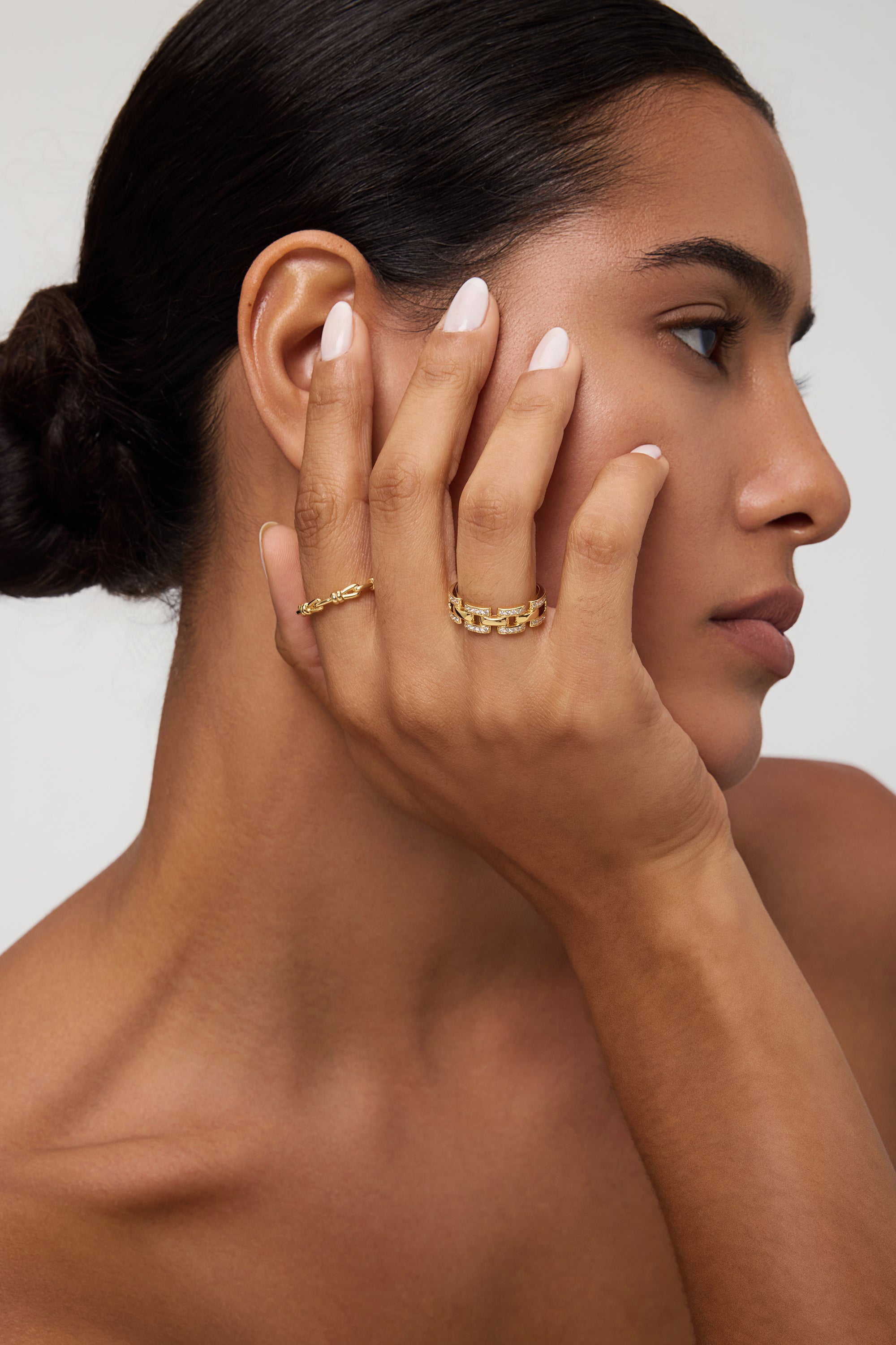 A woman with smooth skin and dark hair in a low bun touches her face, displaying the Noa Link Ring in Gold on her finger. She has natural makeup, nude-colored nails, and gazes to the side against a neutral background.
