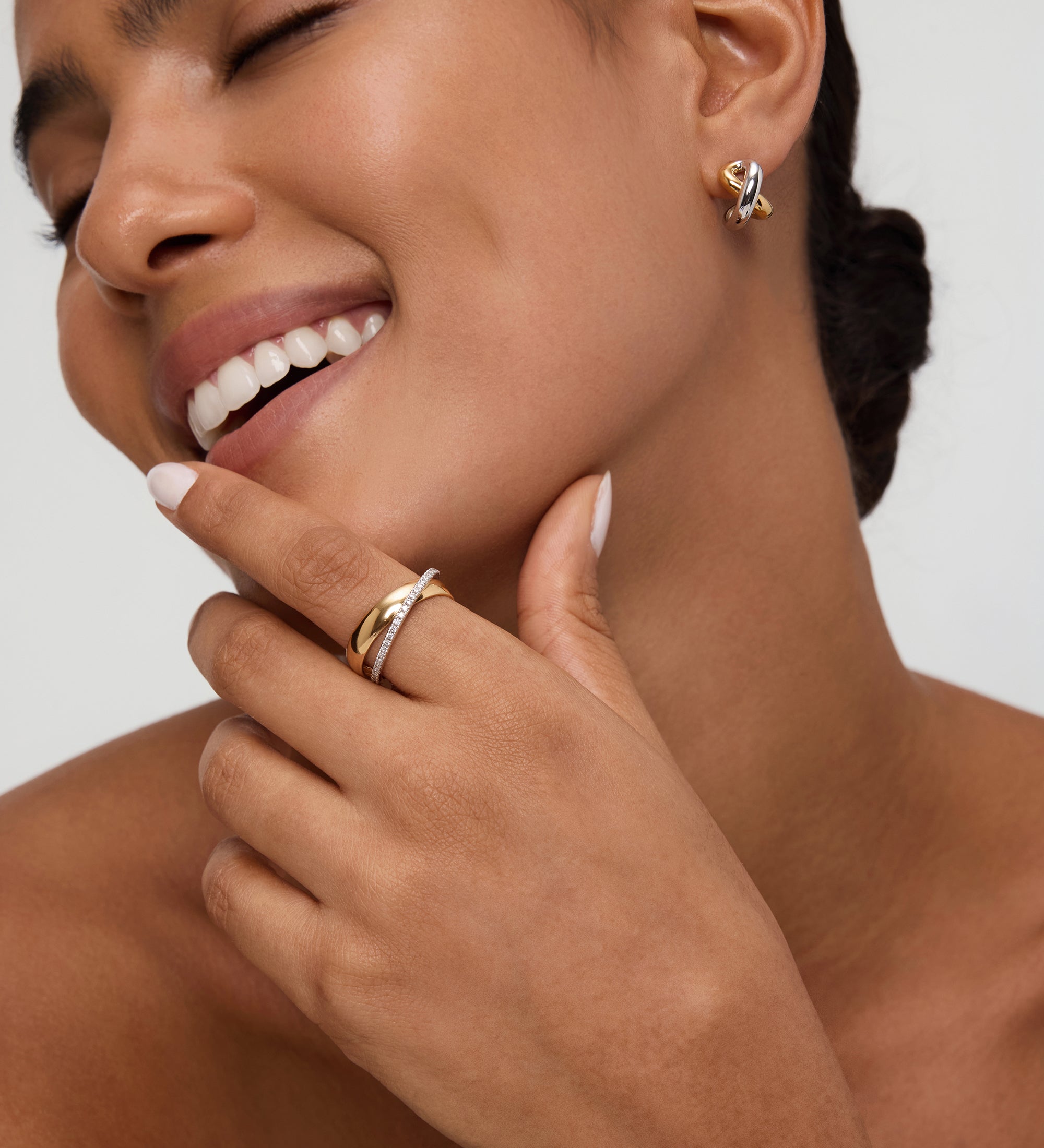 A smiling woman with smooth skin wears the Mia Two-tone Mixed Metal Hoops. She gently touches her chin, highlighting her everyday jewellery and natural beauty, with her hair pulled back and eyes closed.