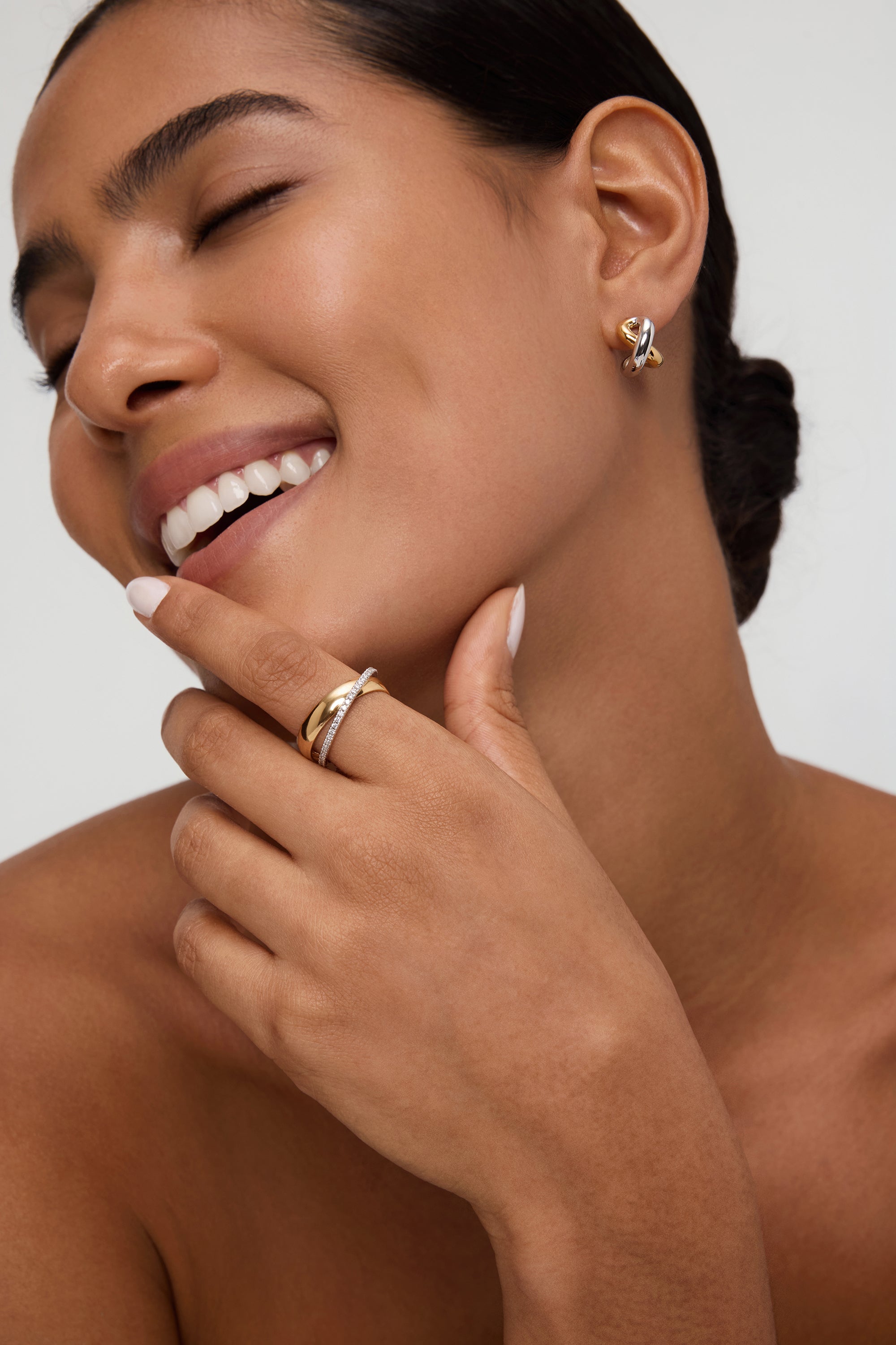 A smiling woman with smooth skin wears the Mia Two-tone Mixed Metal Hoops. She gently touches her chin, highlighting her everyday jewellery and natural beauty, with her hair pulled back and eyes closed.