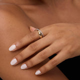 A close-up of a person’s hand with neatly manicured nails, gently resting on their shoulder, showcases the Mixed Metal Interlocking Ring—a gold and diamond design. The background is neutral, and the upper body is partially visible.