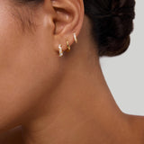 A close-up of a woman's ear with dark hair in a low bun, wearing three small gold huggie earrings, including the Nori Twisted Crystal Huggies in Gold. The background is plain and neutral.