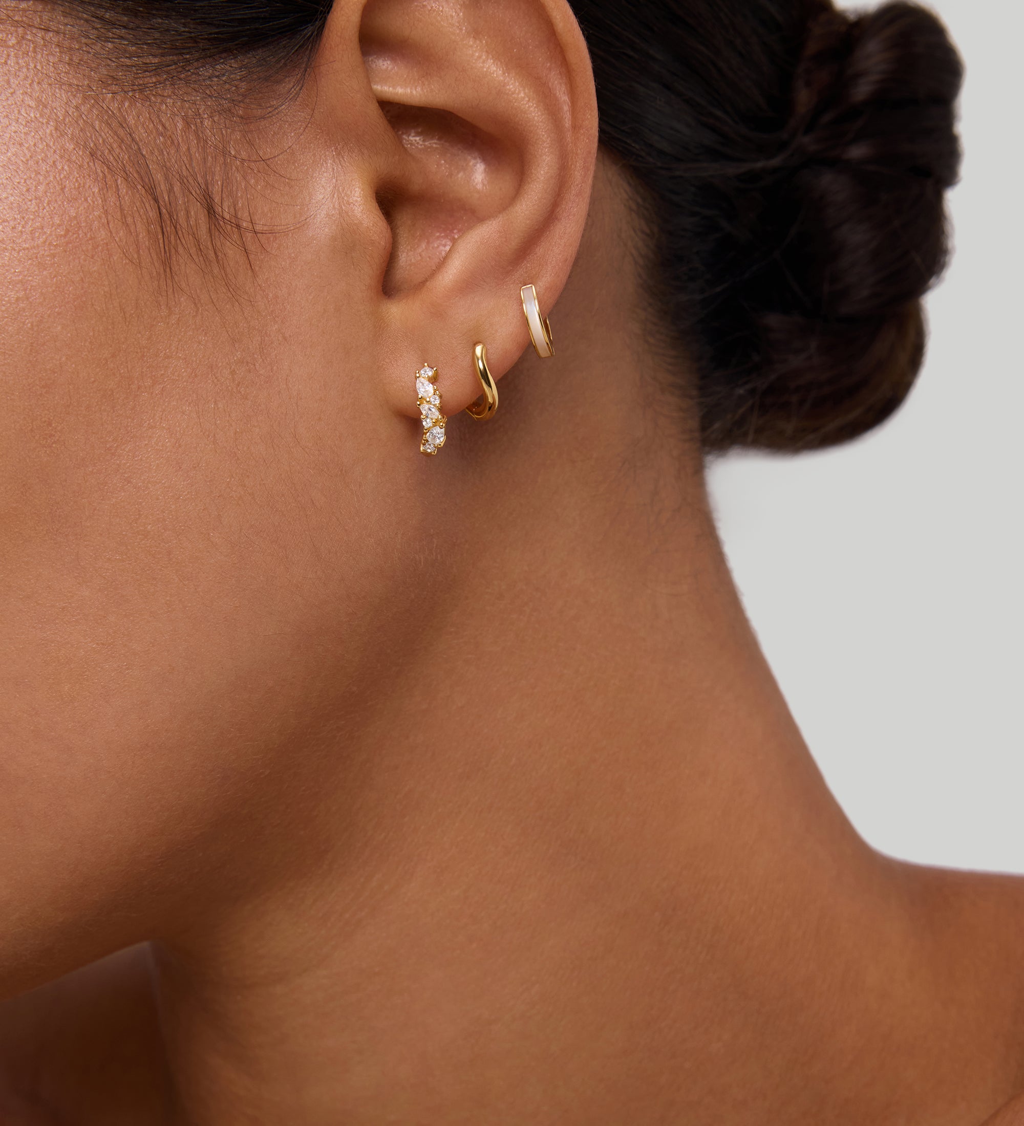 A close-up of a woman's ear with dark hair in a low bun, wearing three small gold huggie earrings, including the Nori Twisted Crystal Huggies in Gold. The background is plain and neutral.