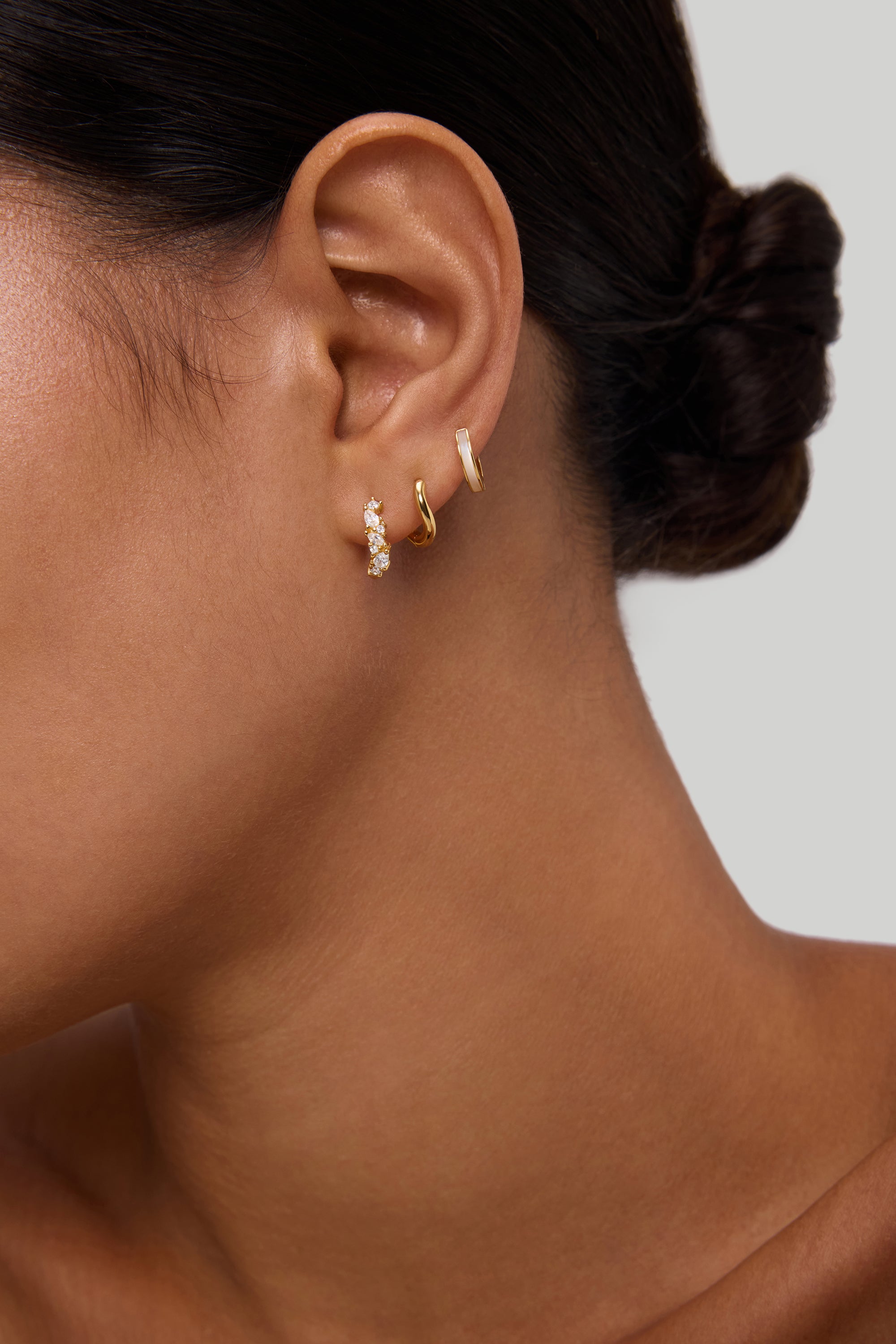 A close-up of a woman's ear with dark hair in a low bun, wearing three small gold huggie earrings, including the Nori Twisted Crystal Huggies in Gold. The background is plain and neutral.