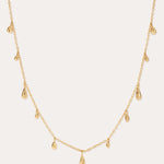 The Rhea Molten Cascade Necklace in Gold features small, evenly spaced teardrop-shaped charms with a subtle hammered texture, all crafted in 14k gold plating and delicately suspended along a fine chain, displayed against a light background.