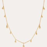 The Rhea Molten Cascade Necklace in Gold features small, evenly spaced teardrop-shaped charms with a subtle hammered texture, all crafted in 14k gold plating and delicately suspended along a fine chain, displayed against a light background.