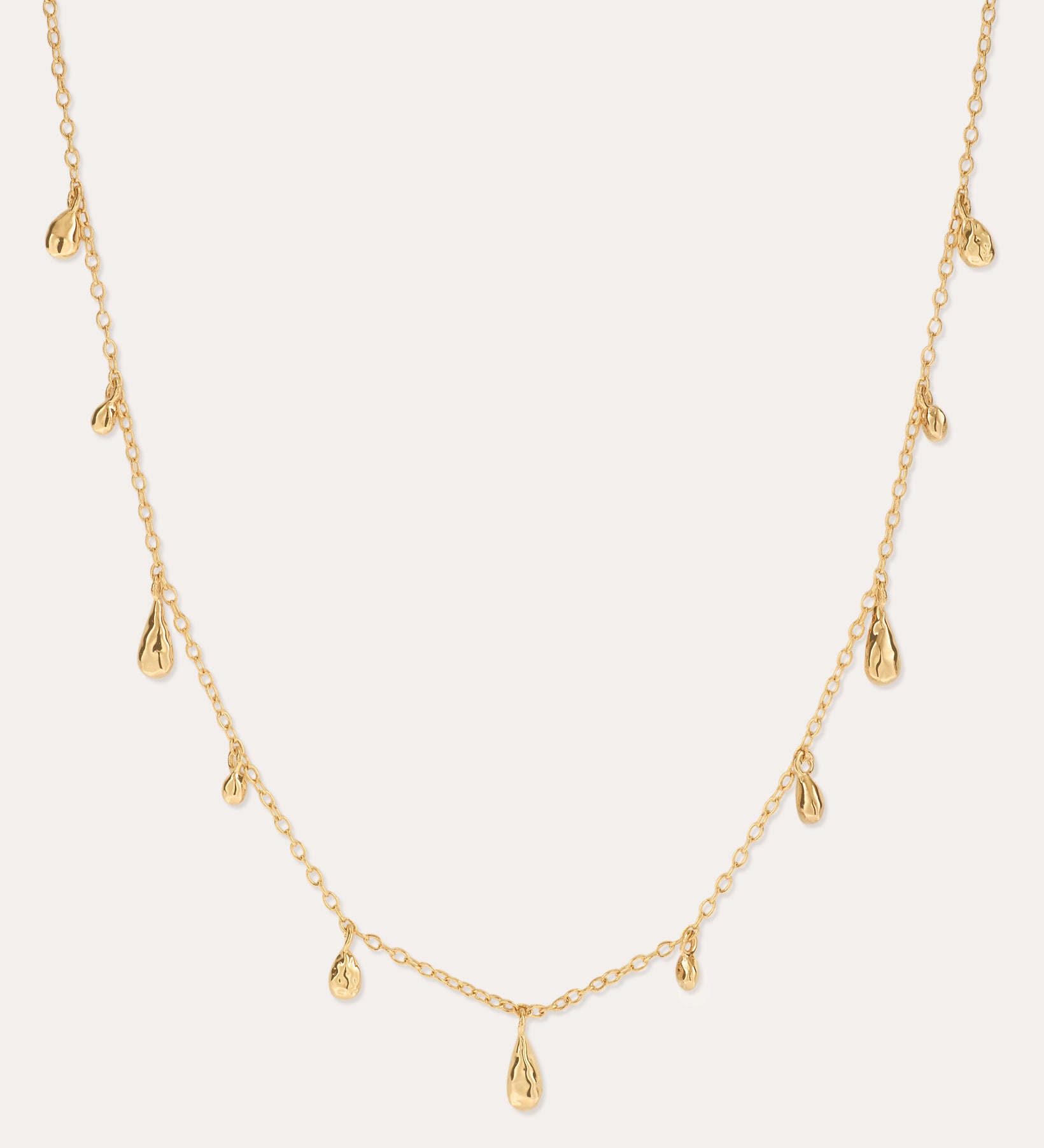The Rhea Molten Cascade Necklace in Gold features small, evenly spaced teardrop-shaped charms with a subtle hammered texture, all crafted in 14k gold plating and delicately suspended along a fine chain, displayed against a light background.