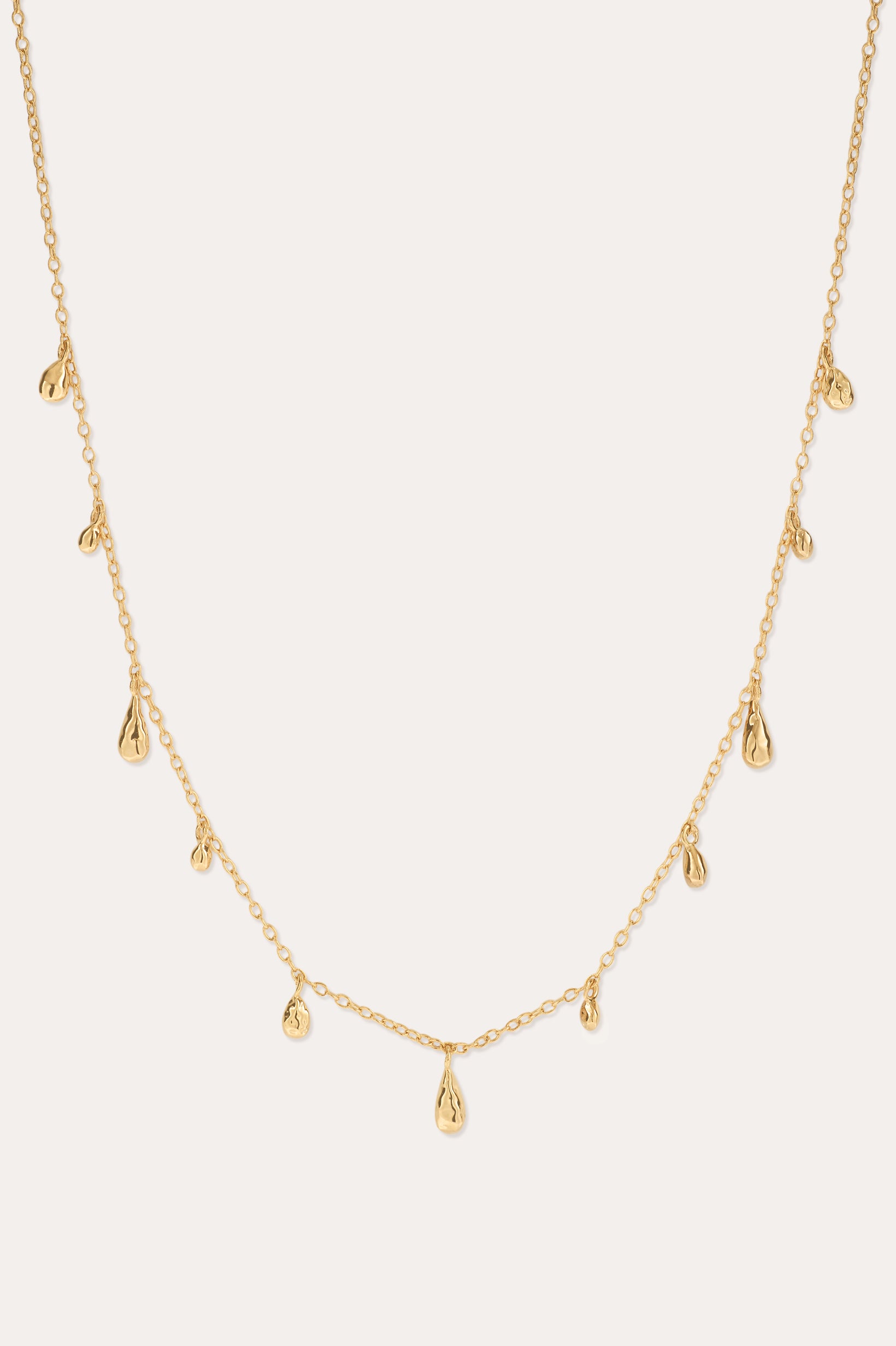 The Rhea Molten Cascade Necklace in Gold features small, evenly spaced teardrop-shaped charms with a subtle hammered texture, all crafted in 14k gold plating and delicately suspended along a fine chain, displayed against a light background.