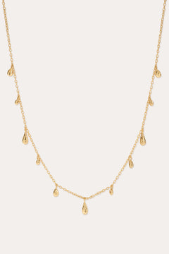 The Rhea Molten Cascade Necklace in Gold features small, evenly spaced teardrop-shaped charms with a subtle hammered texture, all crafted in 14k gold plating and delicately suspended along a fine chain, displayed against a light background.