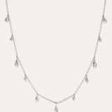 The Rhea Molten Cascade Necklace in Silver features small, teardrop-shaped charms with a subtle hammered texture, crafted from rhodium-plated metal and evenly spaced along a fine chain, displayed against a white background.