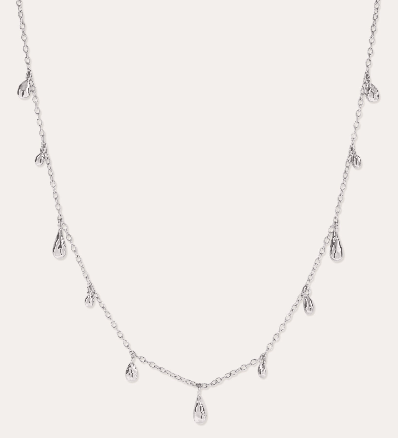 The Rhea Molten Cascade Necklace in Silver features small, teardrop-shaped charms with a subtle hammered texture, crafted from rhodium-plated metal and evenly spaced along a fine chain, displayed against a white background.