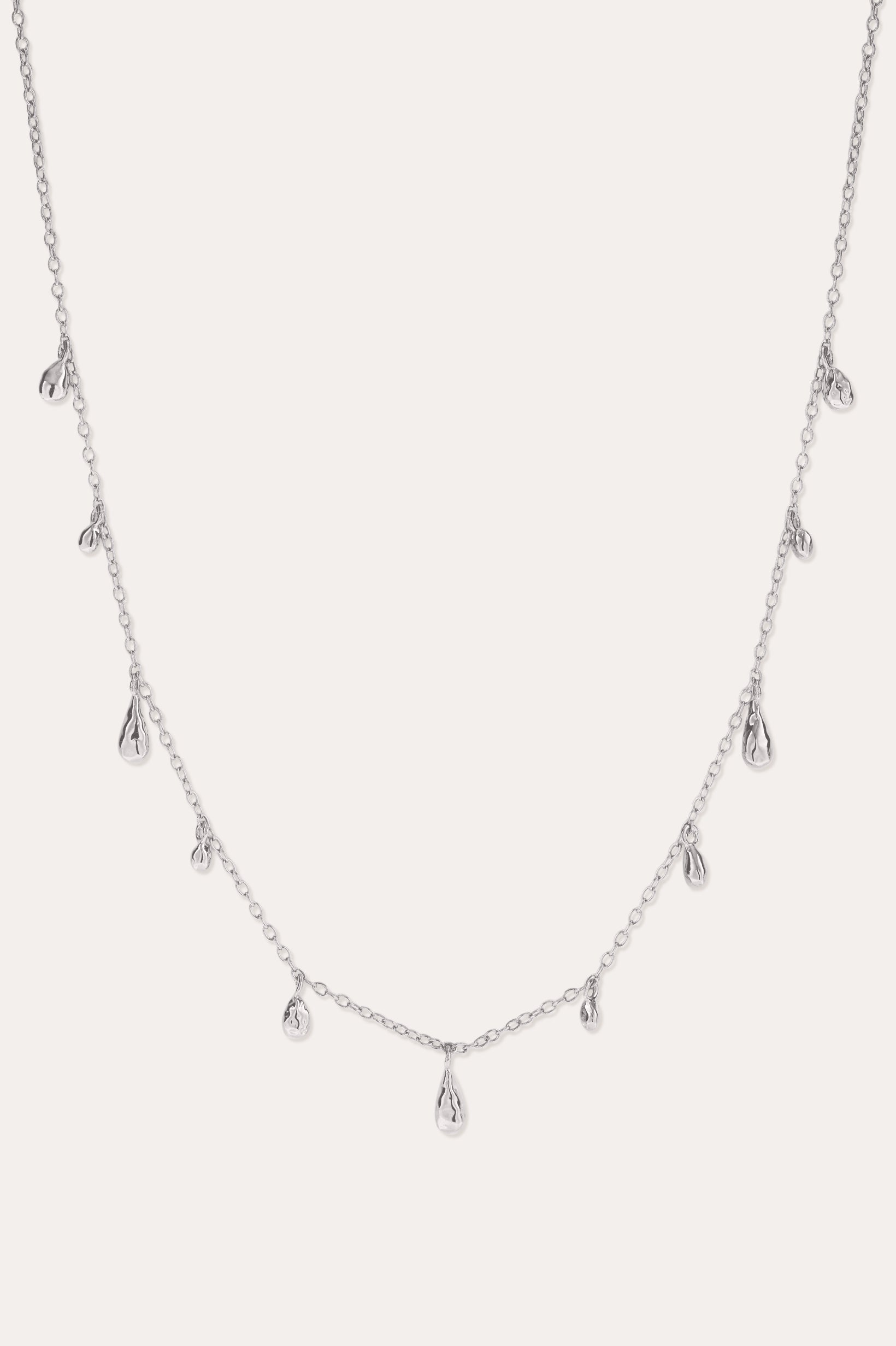 The Rhea Molten Cascade Necklace in Silver features small, teardrop-shaped charms with a subtle hammered texture, crafted from rhodium-plated metal and evenly spaced along a fine chain, displayed against a white background.