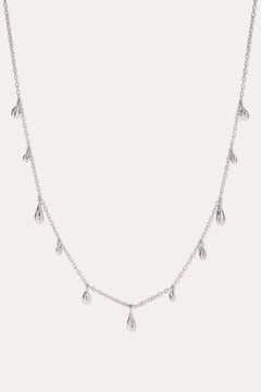 The Rhea Molten Cascade Necklace in Silver features small, teardrop-shaped charms with a subtle hammered texture, crafted from rhodium-plated metal and evenly spaced along a fine chain, displayed against a white background.