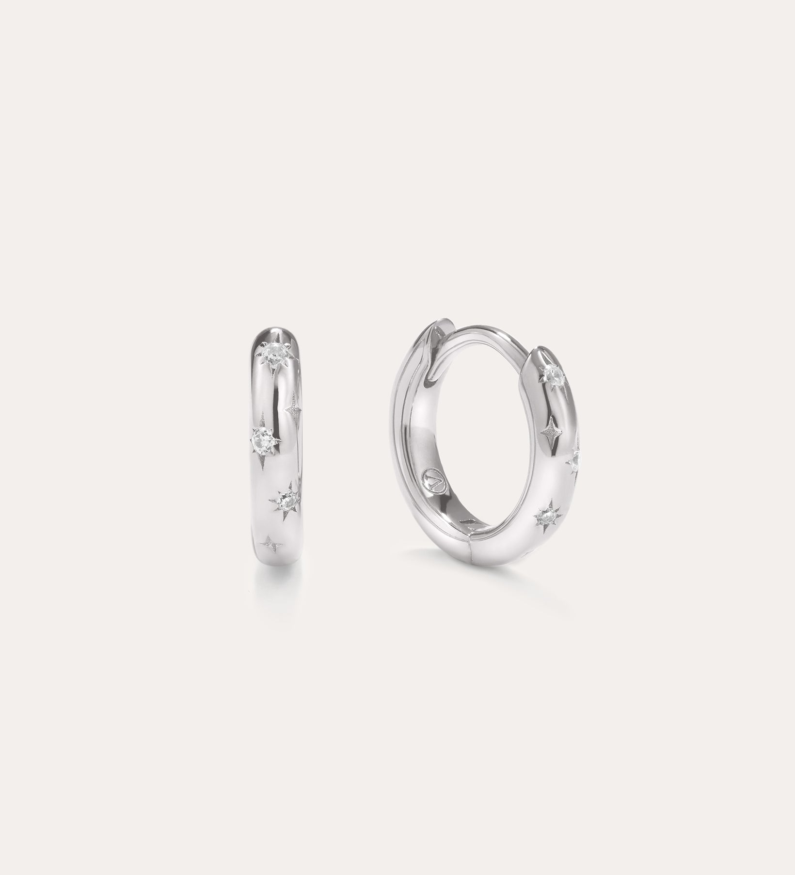 A pair of Starfall Huggies in Silver are displayed against a plain, light background. Crafted in sterling silver, these earrings feature engraved star-shaped designs accented with sparkling cubic zirconia stones, creating a delicate celestial style reminiscent of star burst hoops. The glittering details add an extra touch of brilliance to these elegant pieces.