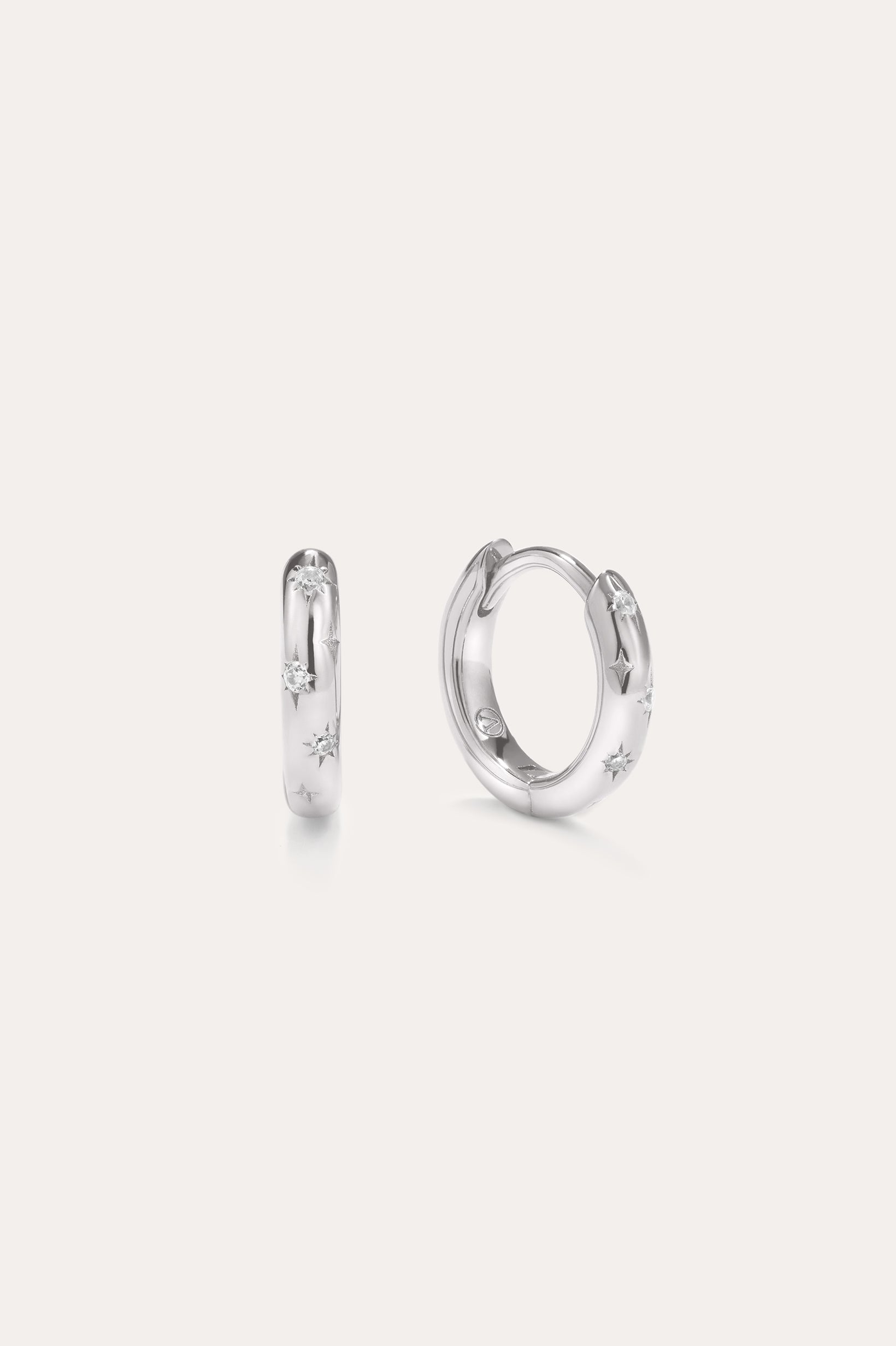 A pair of Starfall Huggies in Silver are displayed against a plain, light background. Crafted in sterling silver, these earrings feature engraved star-shaped designs accented with sparkling cubic zirconia stones, creating a delicate celestial style reminiscent of star burst hoops. The glittering details add an extra touch of brilliance to these elegant pieces.