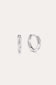 A pair of Starfall Huggies in Silver are displayed against a plain, light background. Crafted in sterling silver, these earrings feature engraved star-shaped designs accented with sparkling cubic zirconia stones, creating a delicate celestial style reminiscent of star burst hoops. The glittering details add an extra touch of brilliance to these elegant pieces.