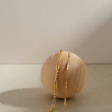 The Carly Paperclip Chain Bead Bracelet in Gold rests gracefully on a smooth, round wooden sphere, highlighted by a neutral beige background with soft lighting and gentle shadows.
