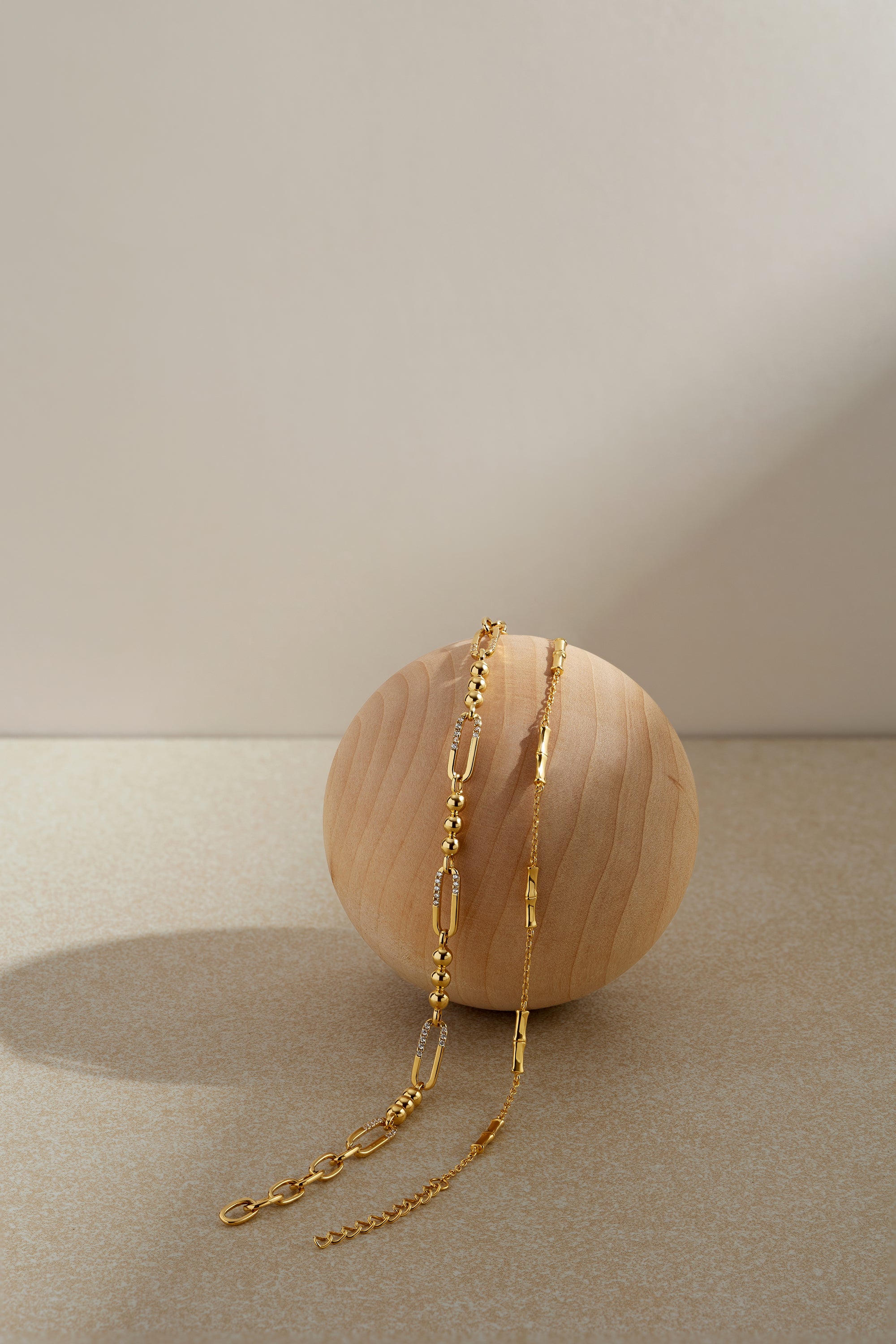The Carly Paperclip Chain Bead Bracelet in Gold rests gracefully on a smooth, round wooden sphere, highlighted by a neutral beige background with soft lighting and gentle shadows.