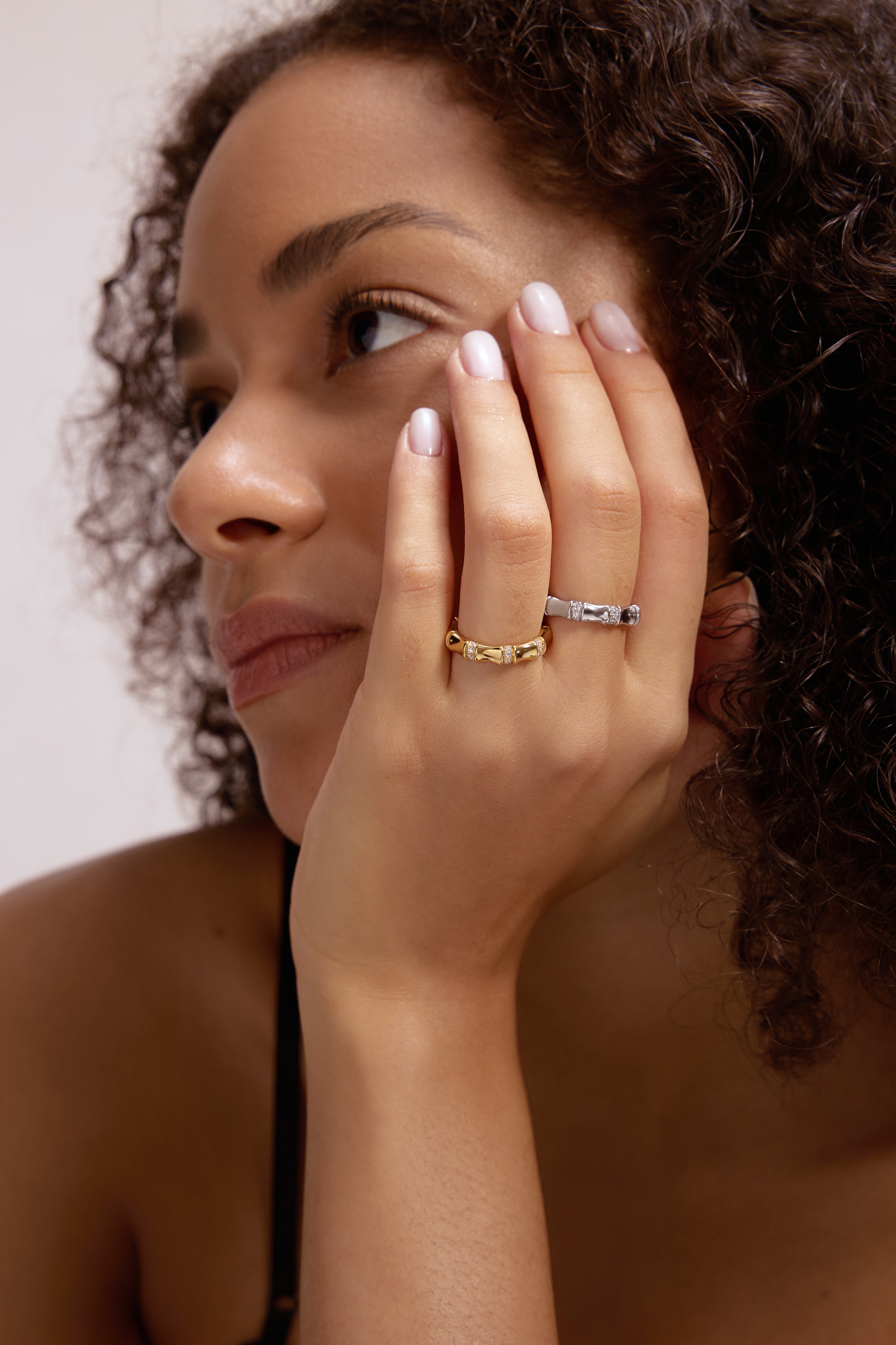 A woman with curly hair, natural makeup, and a thoughtful expression rests her chin on her hand, showcasing the Bamboo Chunky Crystal Ring in Gold.