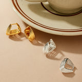 A beige cup and saucer with coffee sit on a light surface. In front, two pairs of modern earrings are displayed, including the Juno Ridged Chunky Hoops in Gold and a pair of silver geometric earrings.