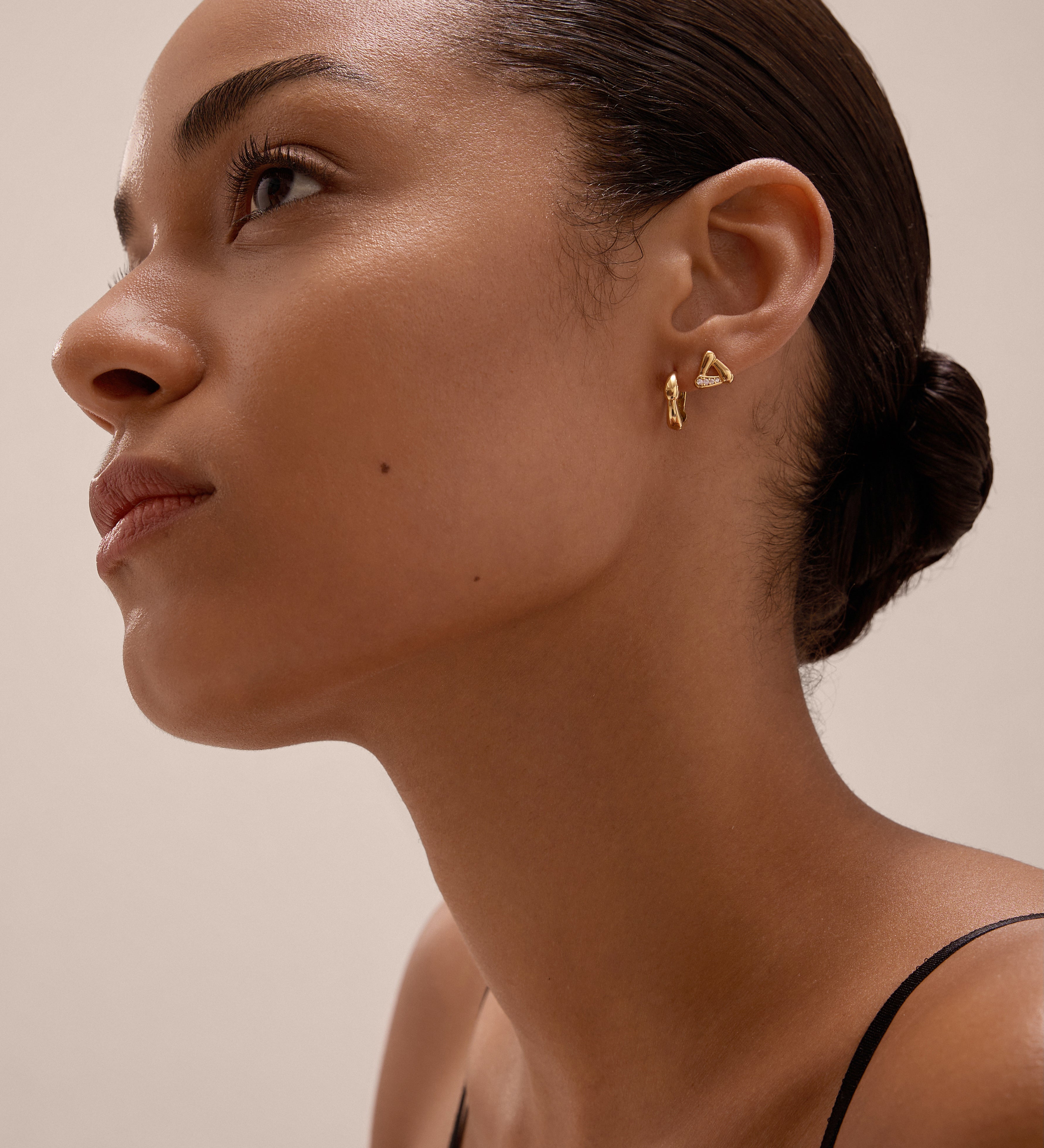 A woman with smooth skin and dark hair in a low bun wears the Colette Droop Link Huggie Earrings in Gold and a black top, gazing upward against a neutral background.