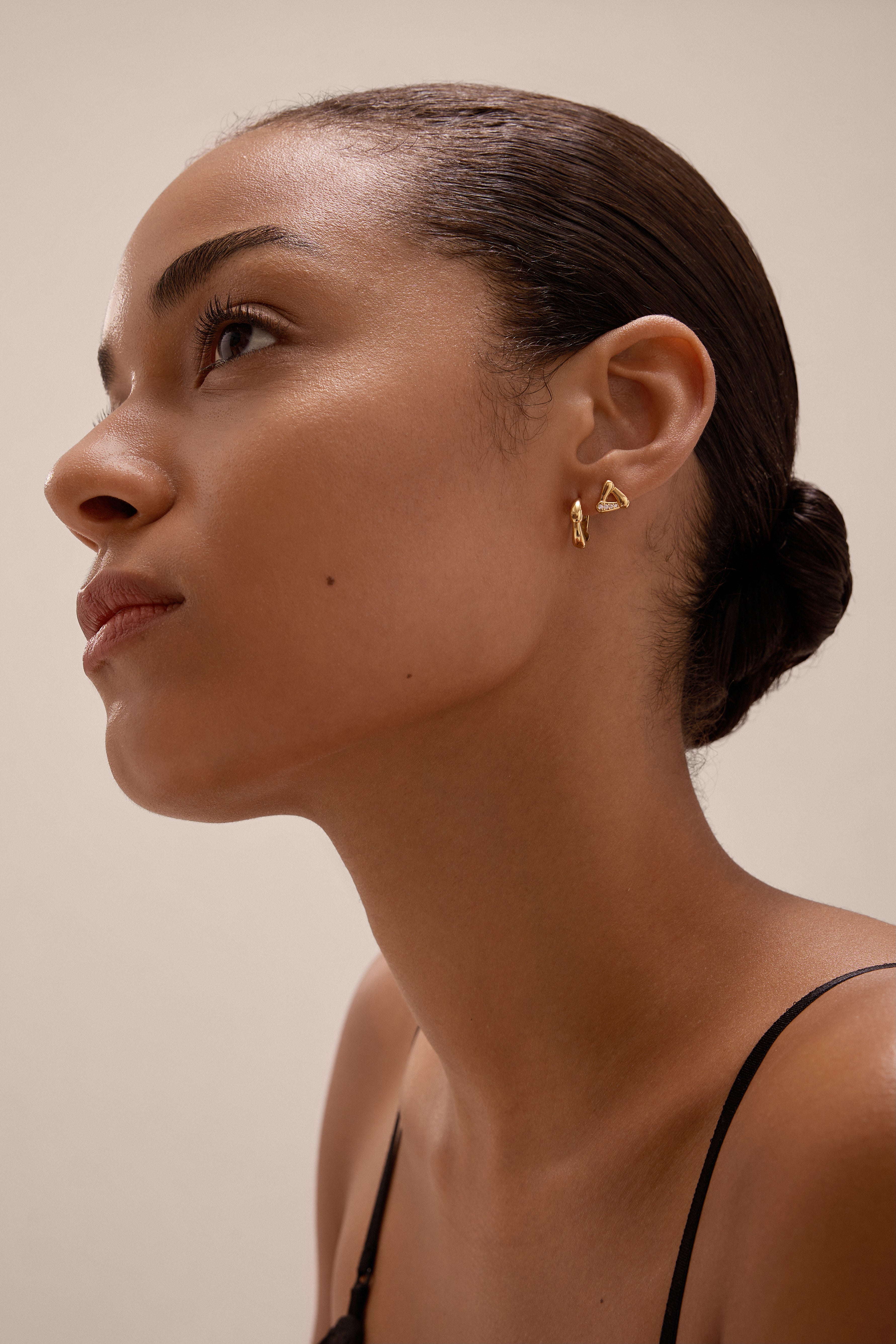 A woman with smooth skin and dark hair in a low bun wears the Colette Droop Link Huggie Earrings in Gold and a black top, gazing upward against a neutral background.