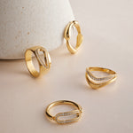 The Colette Link Droop Ring in Gold, featuring an elegant geometric design and small embedded diamonds, is displayed on a light beige surface, with one ring leaning against a textured, cone-shaped object in the background.