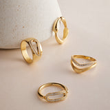 The Colette Link Droop Ring in Gold, featuring an elegant geometric design and small embedded diamonds, is displayed on a light beige surface, with one ring leaning against a textured, cone-shaped object in the background.