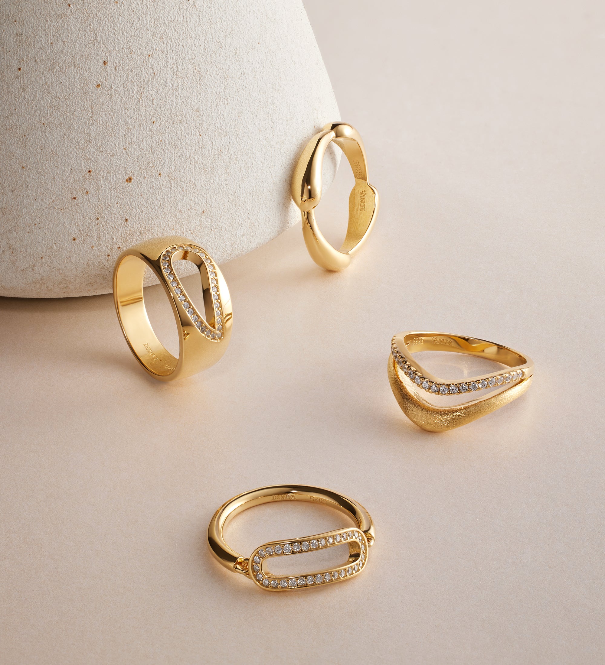 The Colette Link Droop Ring in Gold, featuring an elegant geometric design and small embedded diamonds, is displayed on a light beige surface, with one ring leaning against a textured, cone-shaped object in the background.