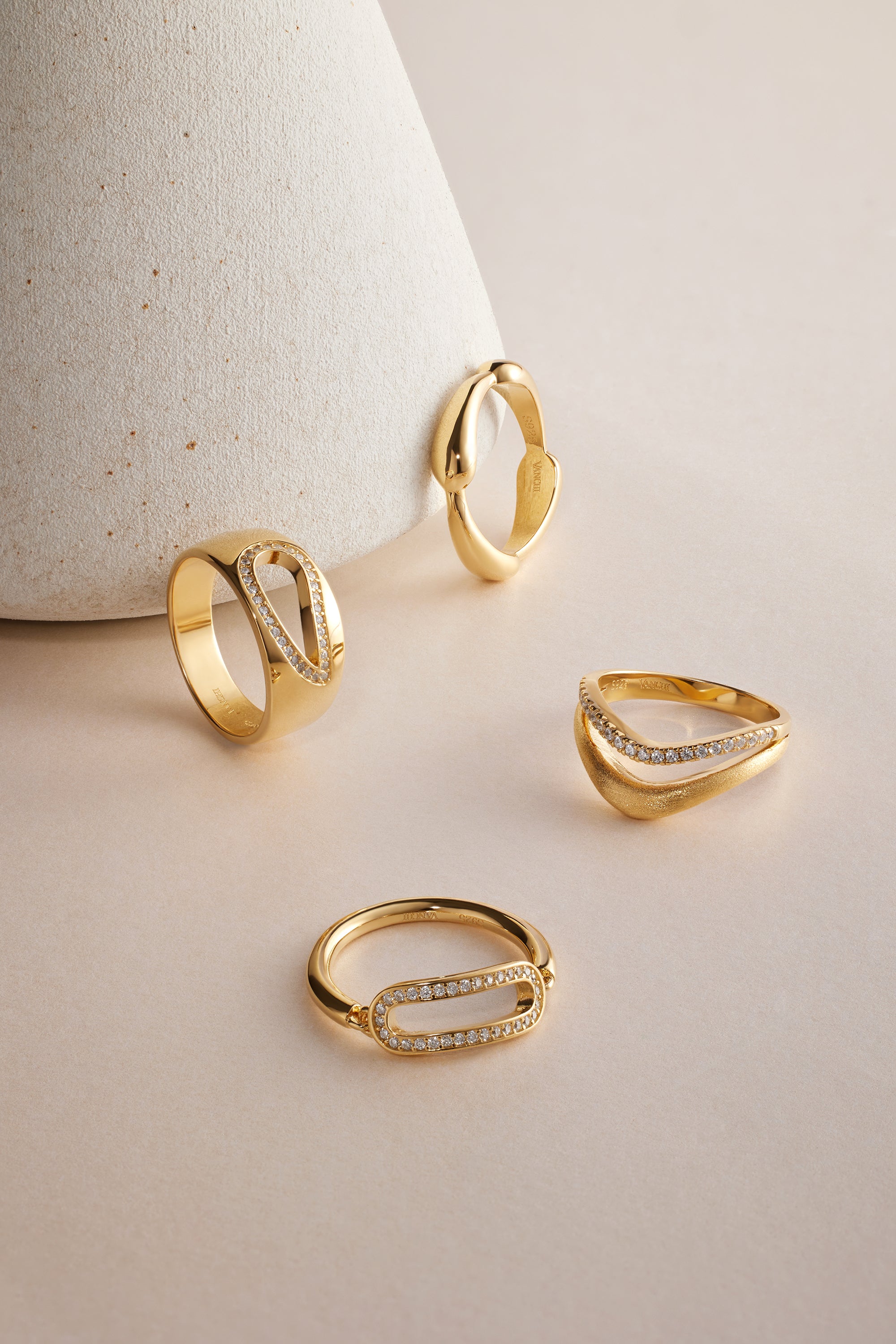 The Colette Link Droop Ring in Gold, featuring an elegant geometric design and small embedded diamonds, is displayed on a light beige surface, with one ring leaning against a textured, cone-shaped object in the background.