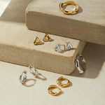 A display of gold and silver jewelry—including statement rings, hoops, studs, and geometric shapes—featuring the Colette Link Droop Ring in Gold on stacked beige stone platforms against a soft, neutral backdrop.