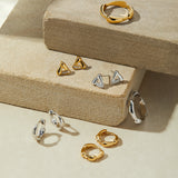 A display of gold and silver jewelry—including statement rings, hoops, studs, and geometric shapes—featuring the Colette Link Droop Ring in Gold on stacked beige stone platforms against a soft, neutral backdrop.