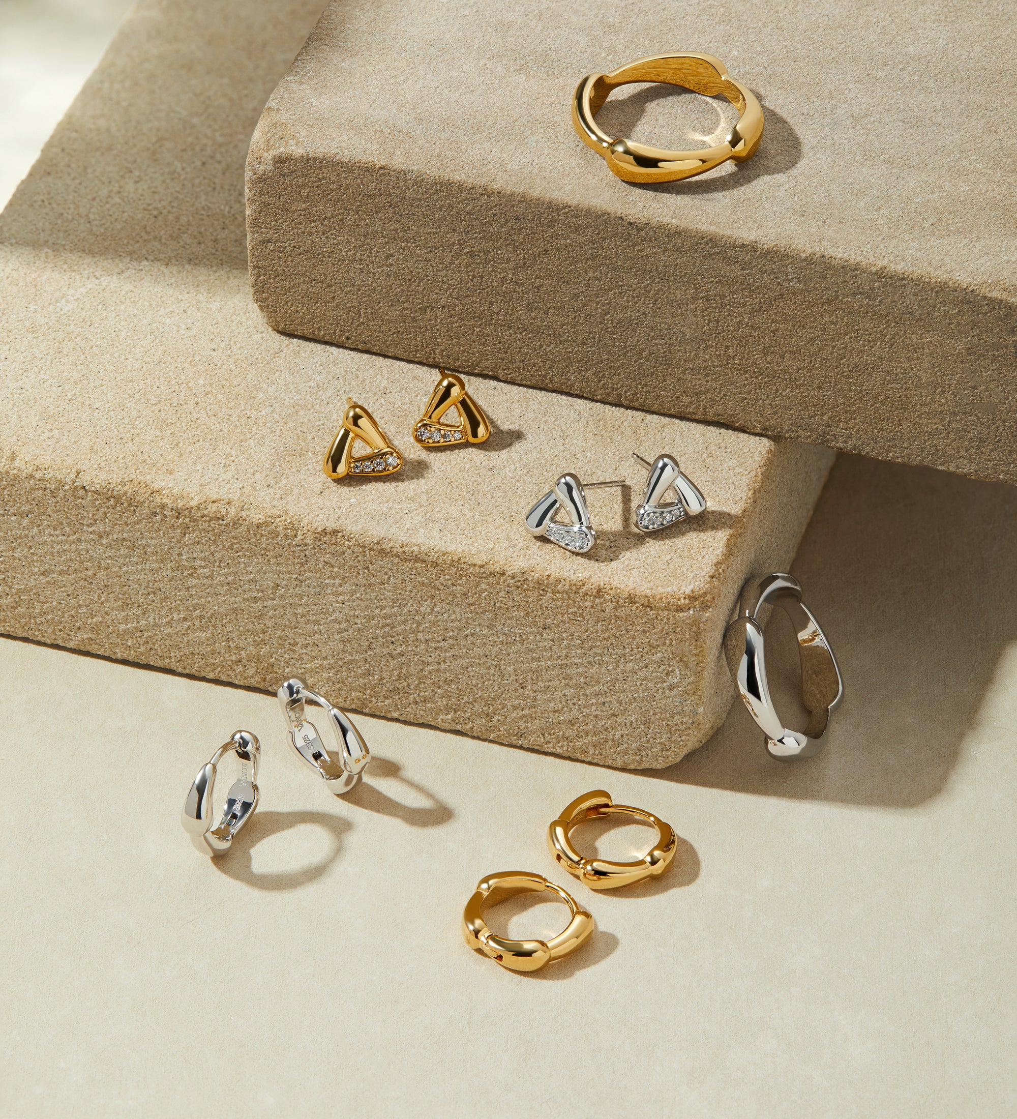 A display of gold and silver jewelry—including statement rings, hoops, studs, and geometric shapes—featuring the Colette Link Droop Ring in Gold on stacked beige stone platforms against a soft, neutral backdrop.