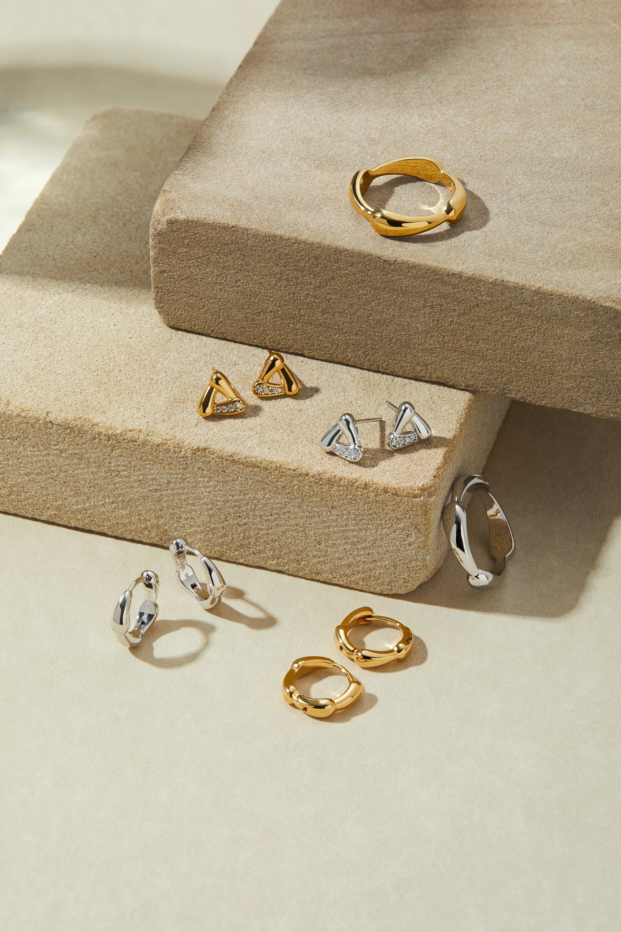 A display of gold and silver jewelry—including statement rings, hoops, studs, and geometric shapes—featuring the Colette Link Droop Ring in Gold on stacked beige stone platforms against a soft, neutral backdrop.