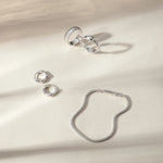 Four silver rings, including the Colette Link Droop Ring in Silver, and a silver bracelet are arranged on a cream-colored surface with soft, natural shadows—an elegant display of skate stopper jewelry.