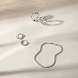 Four silver rings, including the Colette Link Droop Ring in Silver, and a silver bracelet are arranged on a cream-colored surface with soft, natural shadows—an elegant display of skate stopper jewelry.