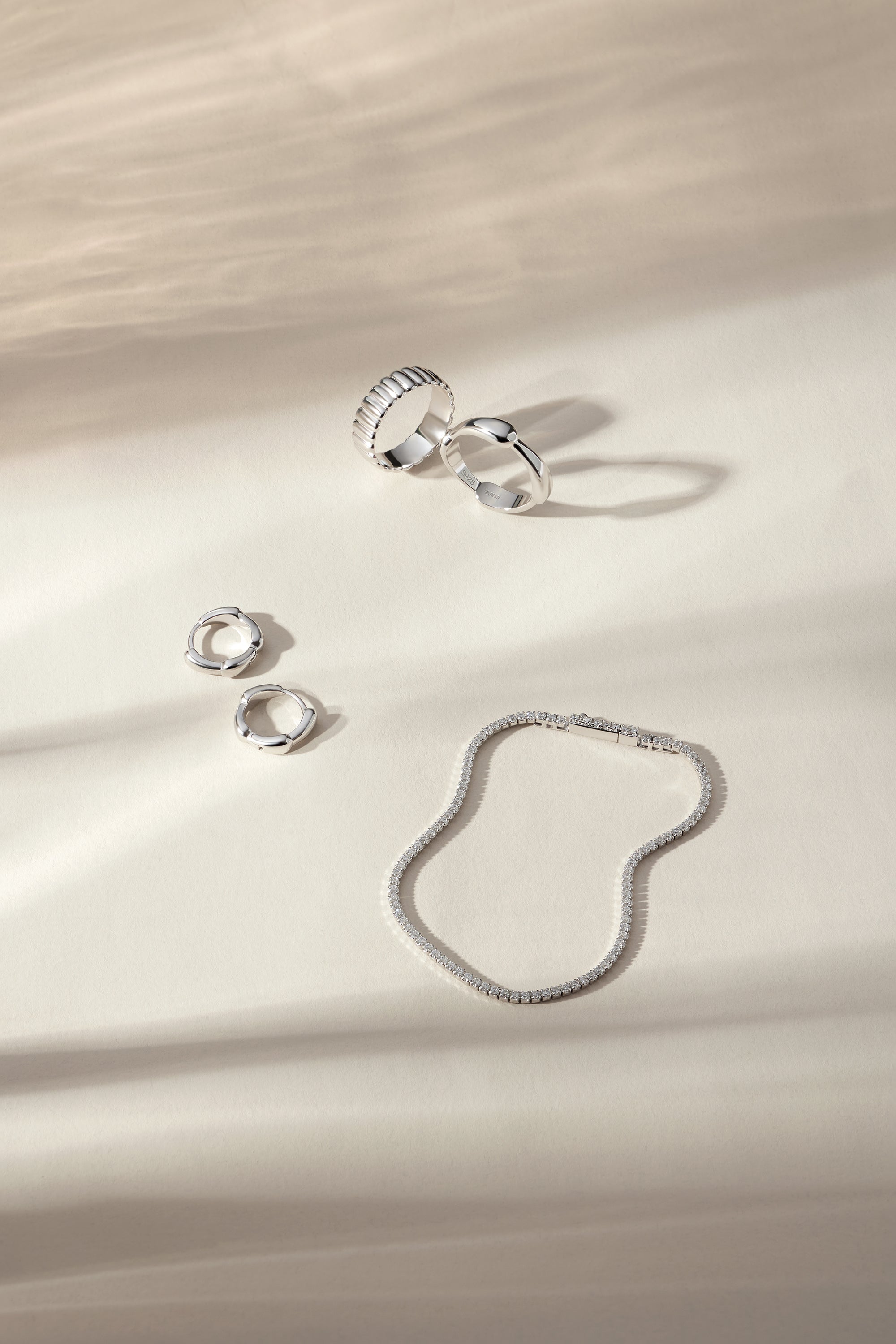 Four silver rings, including the Colette Link Droop Ring in Silver, and a silver bracelet are arranged on a cream-colored surface with soft, natural shadows—an elegant display of skate stopper jewelry.