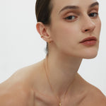 A woman with slicked-back brown hair poses against a plain background, wearing subtle makeup, gold hoop earrings, and the Crescent Moon Crystal Charm Beaded Necklace in Gold. She gazes softly at the camera.