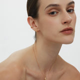 A woman with slicked-back brown hair poses against a plain background, wearing subtle makeup, gold hoop earrings, and the Crescent Moon Crystal Charm Beaded Necklace in Gold. She gazes softly at the camera.