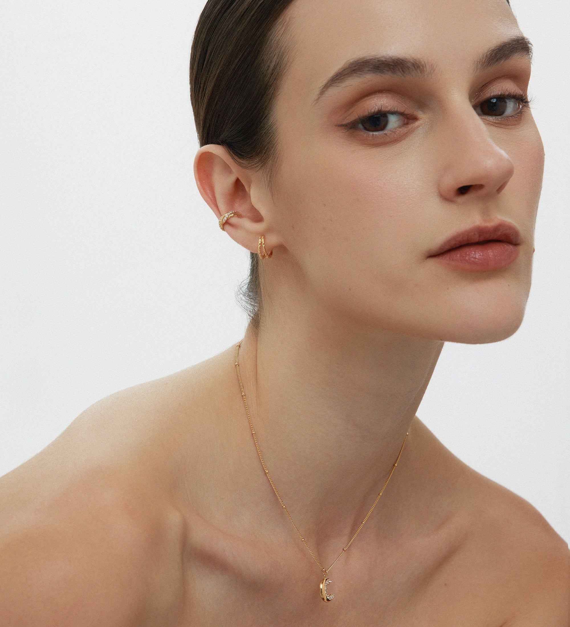 A woman with slicked-back brown hair poses against a plain background, wearing subtle makeup, gold hoop earrings, and the Crescent Moon Crystal Charm Beaded Necklace in Gold. She gazes softly at the camera.