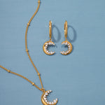 The Crescent Moon Crystal Charm Beaded Necklace in Gold is displayed on a blue background, featuring a crescent moon pendant with sparkling white stones on an elegant beaded chain.