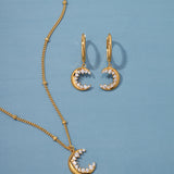 The Crescent Moon Crystal Charm Beaded Necklace in Gold is displayed on a blue background, featuring a crescent moon pendant with sparkling white stones on an elegant beaded chain.