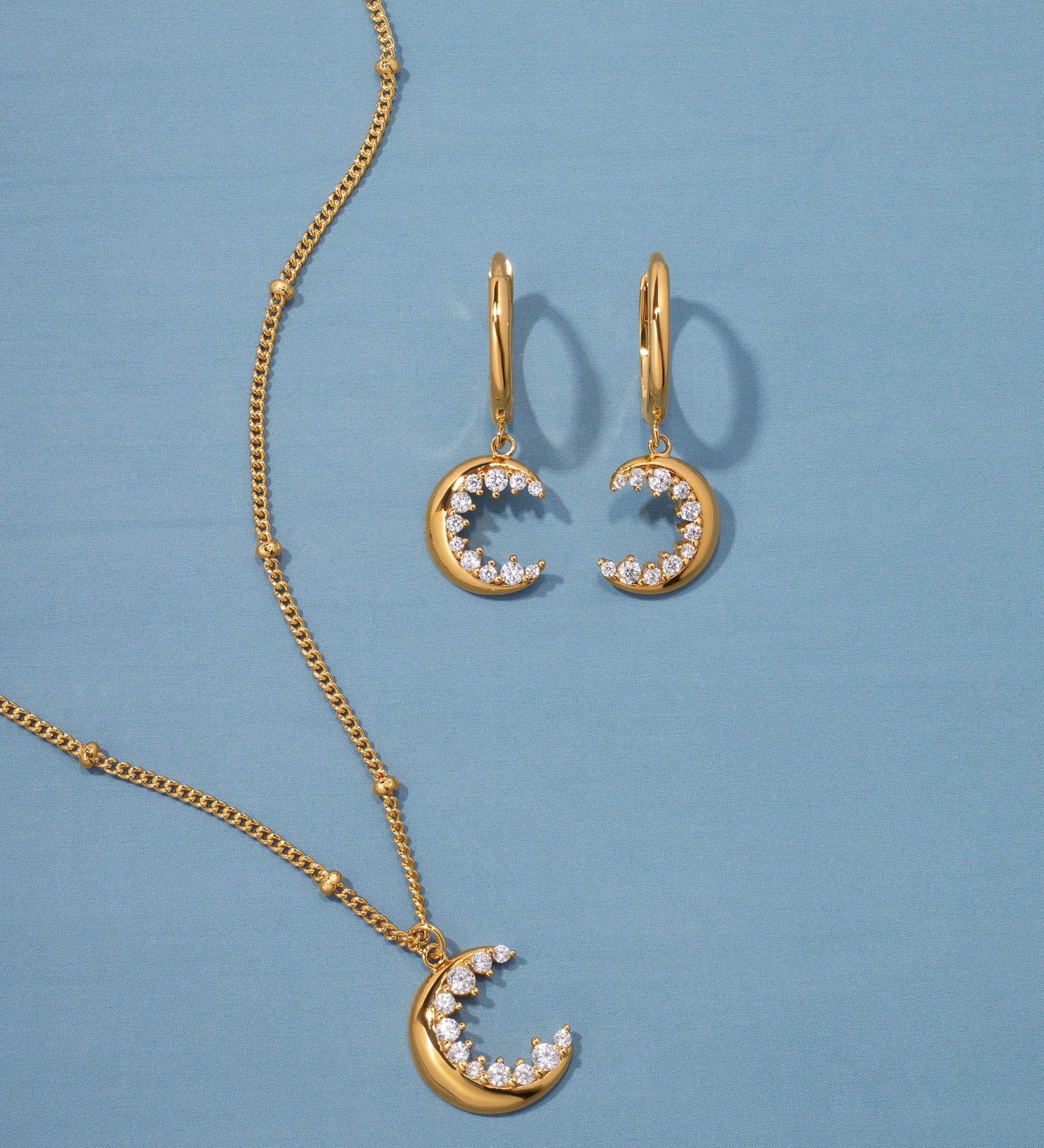 The Crescent Moon Crystal Charm Beaded Necklace in Gold is displayed on a blue background, featuring a crescent moon pendant with sparkling white stones on an elegant beaded chain.