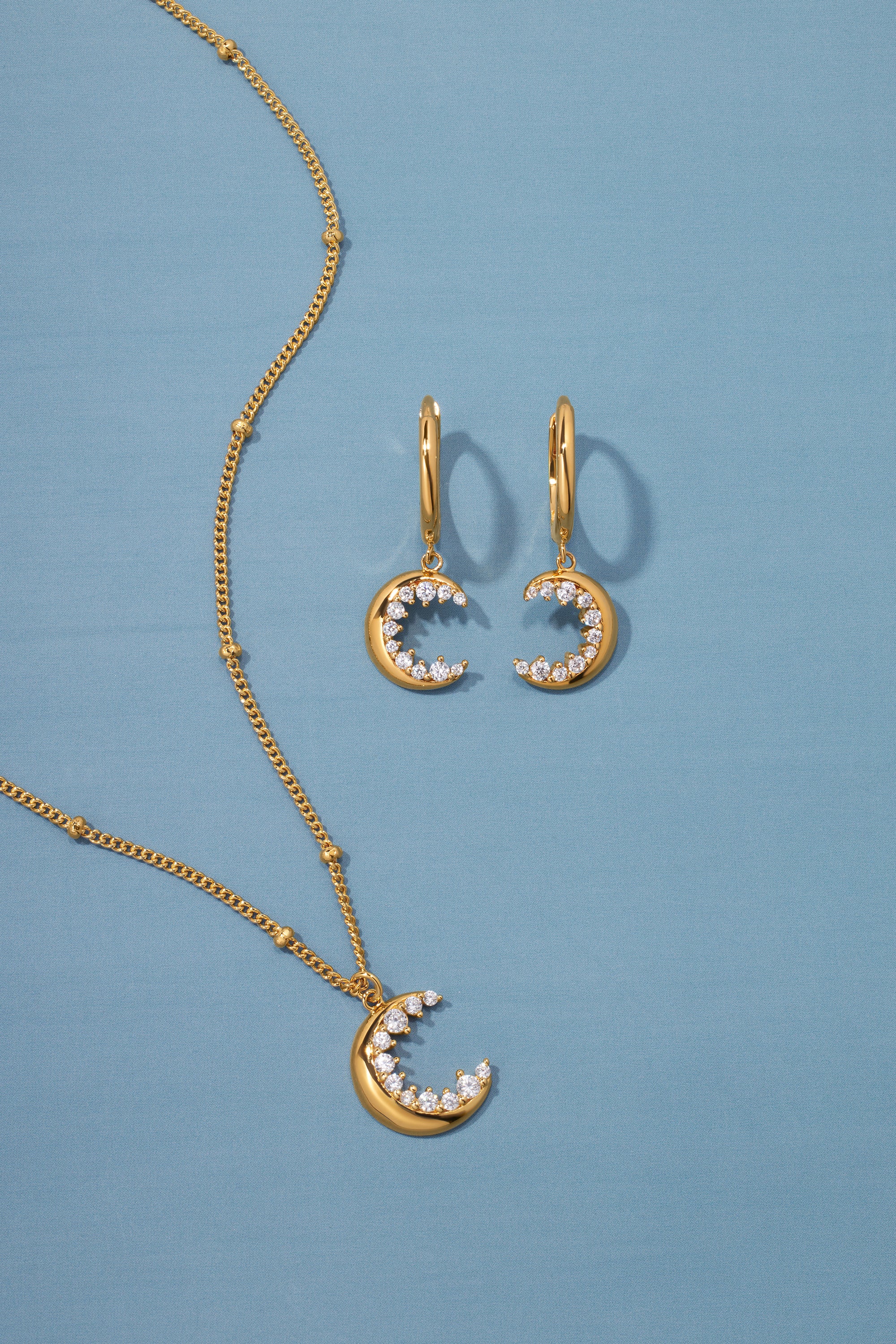 The Crescent Moon Crystal Charm Beaded Necklace in Gold is displayed on a blue background, featuring a crescent moon pendant with sparkling white stones on an elegant beaded chain.
