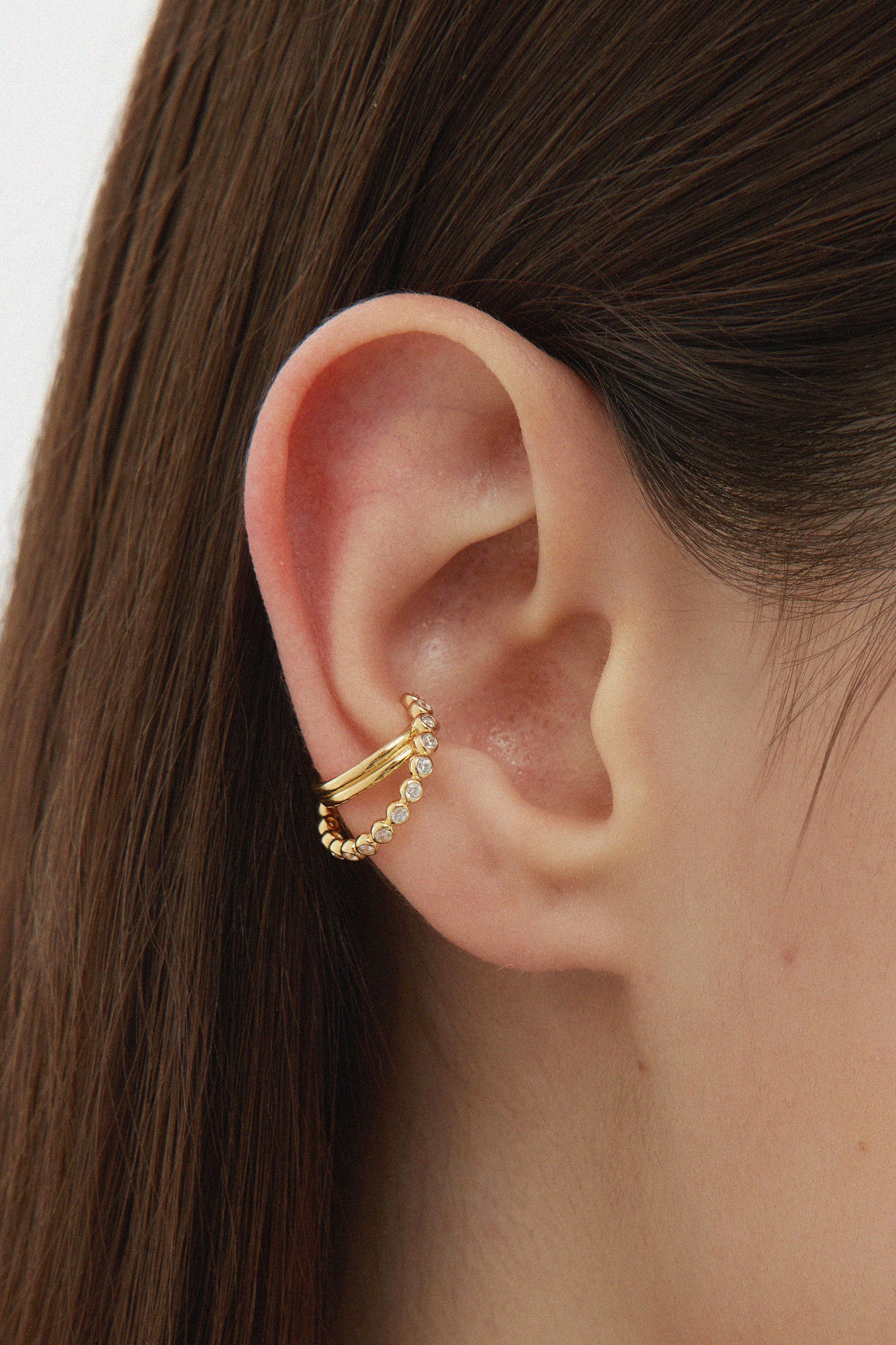 Close-up of a person’s ear with two gold ear cuffs, one smooth and one beaded. The person has straight brown hair and fair skin. Add sparkle by pairing these cuffs with the Vina Crystal Bezel Ear Cuff in Gold.
