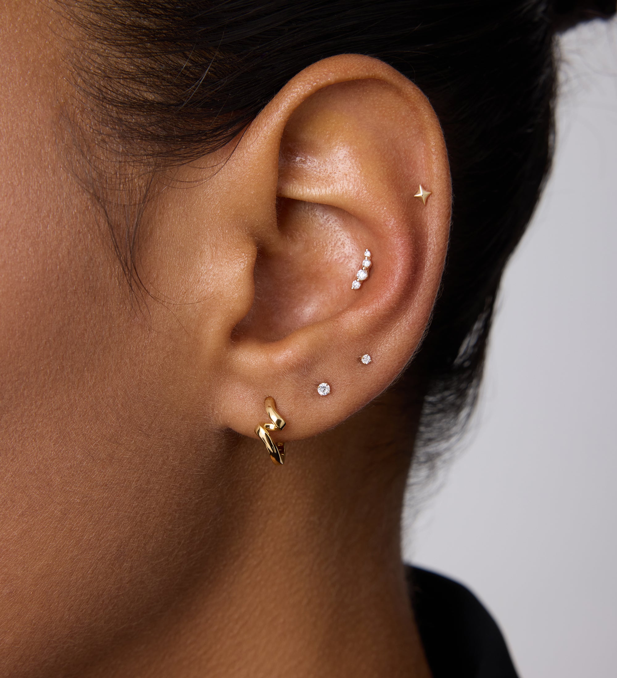 A close-up of a person’s ear with medium brown skin and dark hair wearing multiple piercings, including the 14K Solid White Gold Lab-grown Diamond Prong Set Flat Back Stud - 1.5mm in the lobe alongside gold and silver studs.
