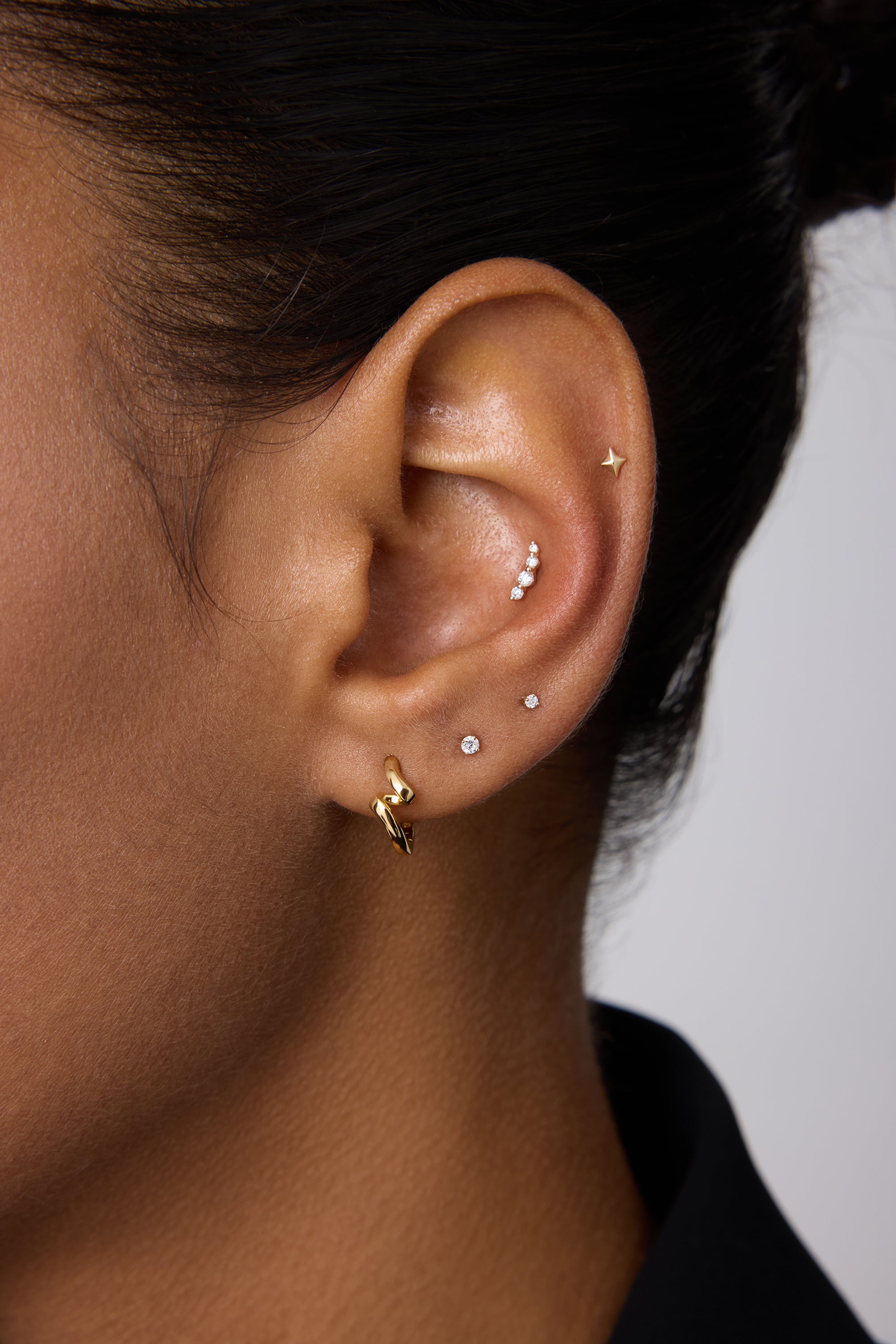 A close-up of a person’s ear with medium brown skin and dark hair wearing multiple piercings, including the 14K Solid White Gold Lab-grown Diamond Prong Set Flat Back Stud - 1.5mm in the lobe alongside gold and silver studs.