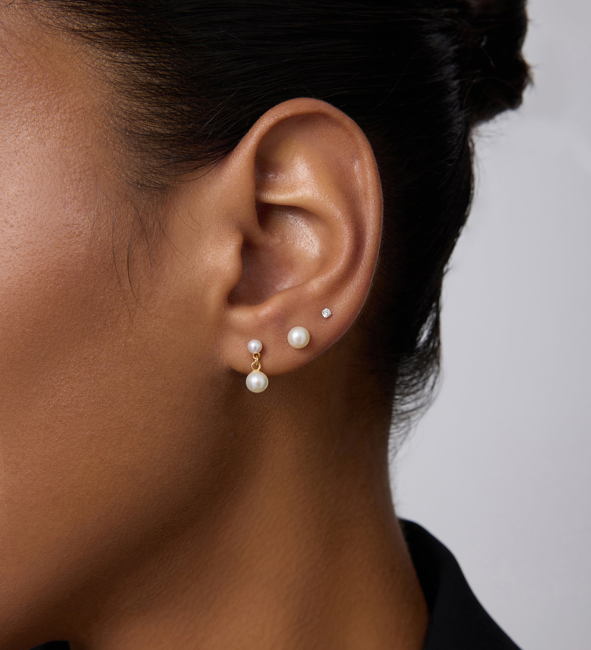 Close-up of a person’s ear wearing three earrings: a 14K solid yellow gold lab-grown diamond prong set flat back stud (2mm), a medium pearl stud, and a gold hoop with dangling pearl. The person has smooth skin and dark hair pulled back.