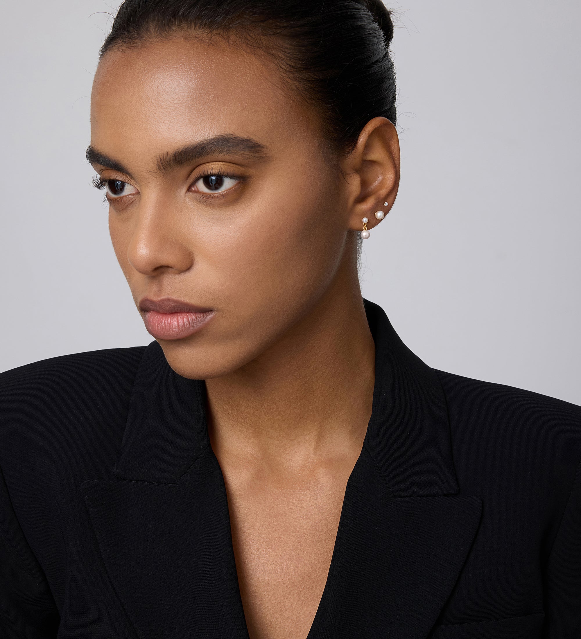 A woman with smooth brown skin and dark hair pulled back wears a black blazer and a 14K solid yellow gold lab-grown diamond prong set flat back stud (2mm), looking slightly to the side against a plain light background.